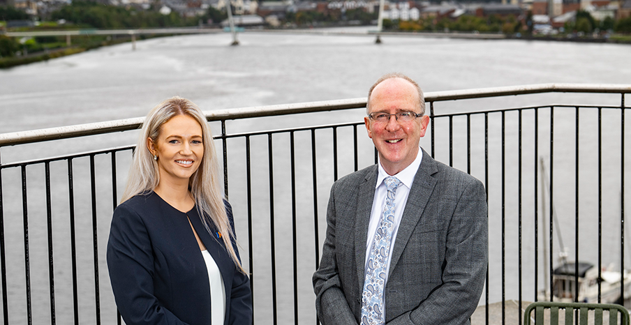 A man and a woman are posing for a photograph on a balcony that overlooks the Peace Bridge 