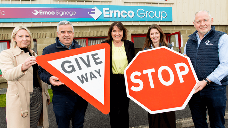 Three women and two men are standing in front of Ernco Group's office and holding road signs